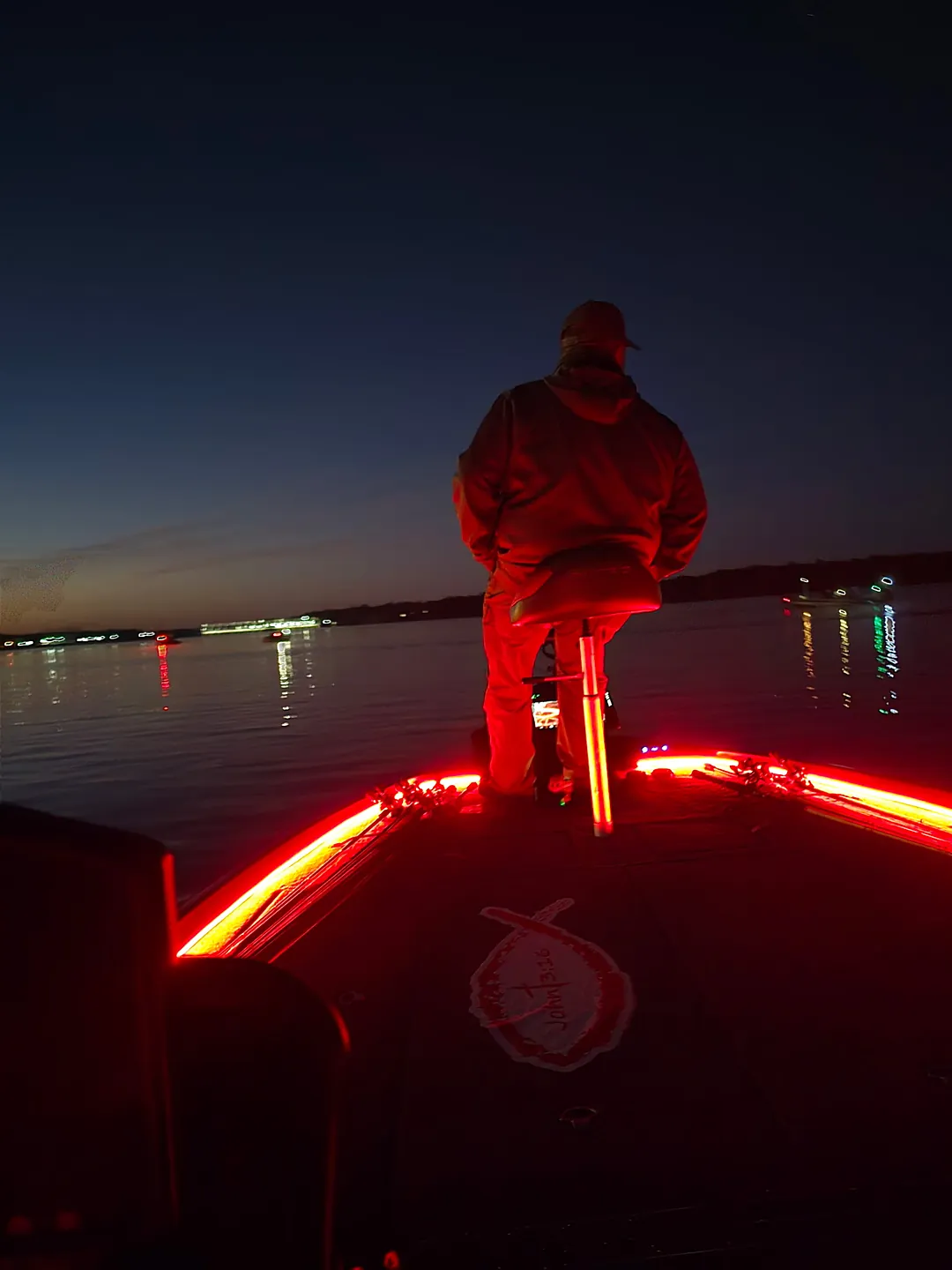 An angler standing on the bow before daylight, lit red by the boat's deck lights, other boats' lights on the water